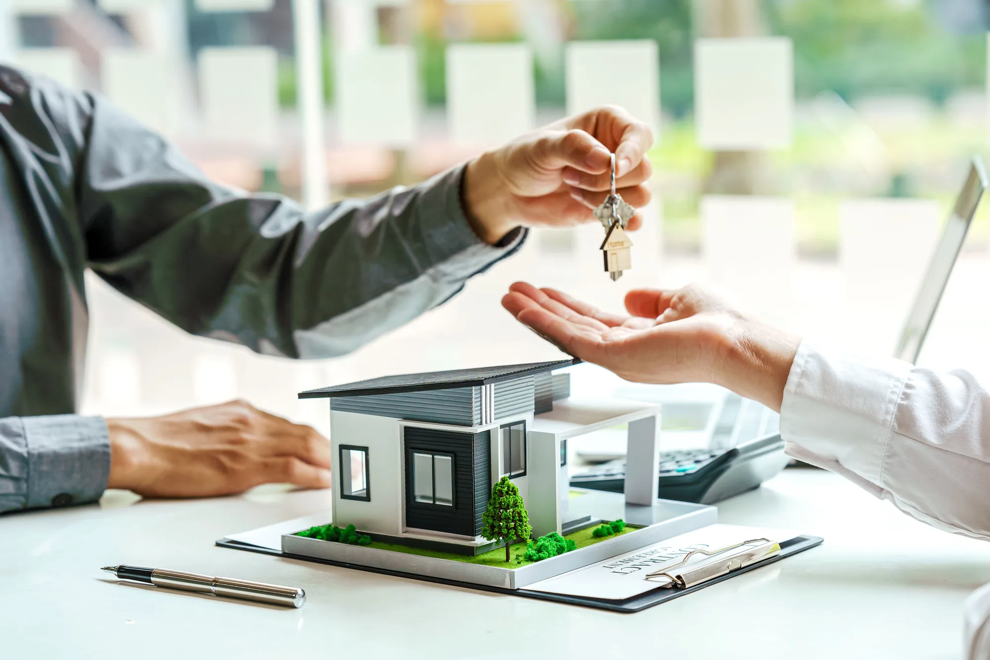 Un agente inmobiliario entregando las llaves de una casa sobre un escritorio con una maqueta y un contrato